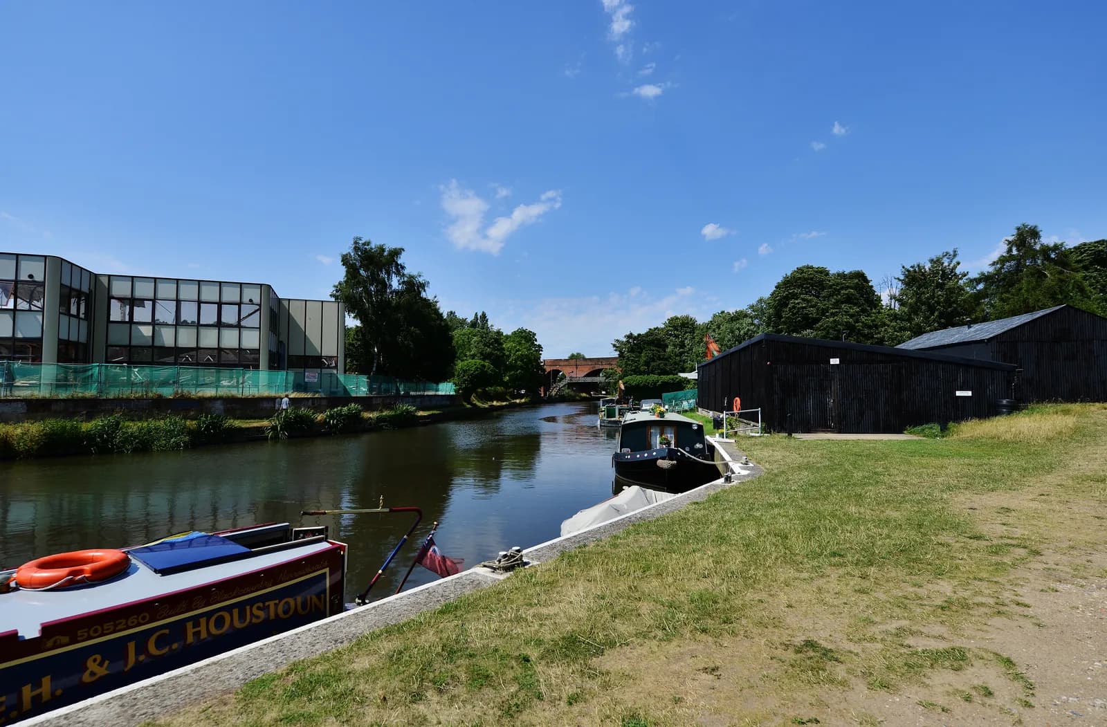 A canal with several boats docked, modern buildings on the left bank, and black wooden structures with grassy grounds on the right bank under a clear blue sky.