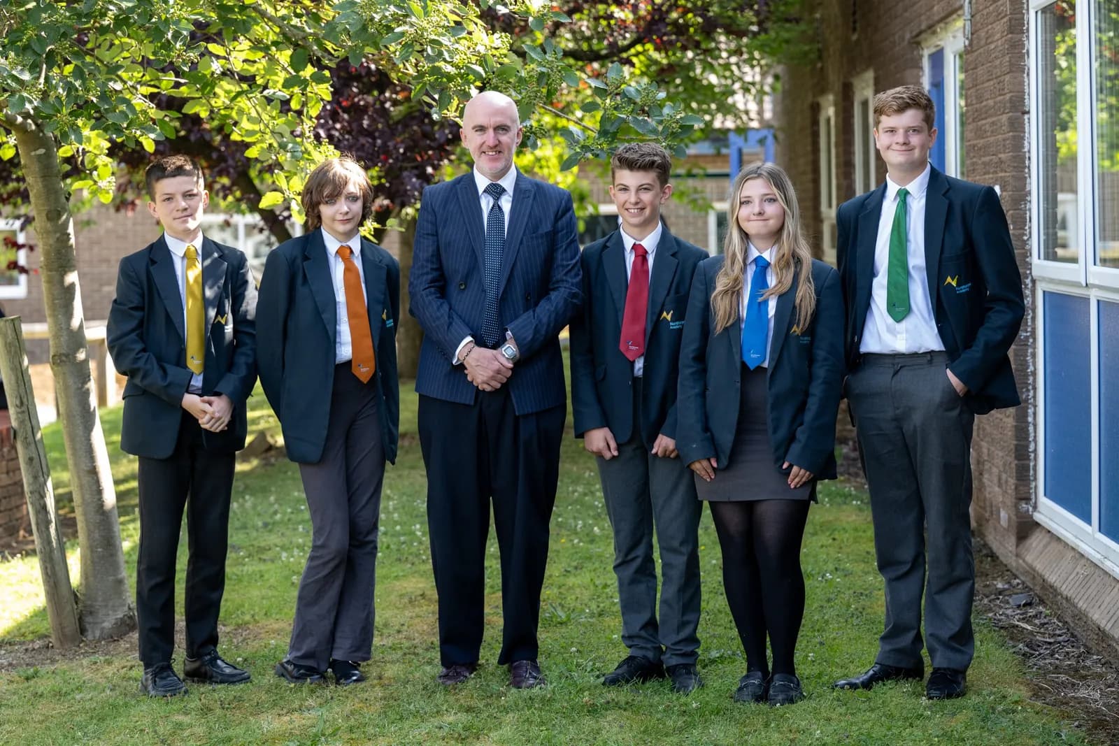 A man in a suit stands with five teenagers in school blazers and ties on a grassy patch outside a building.