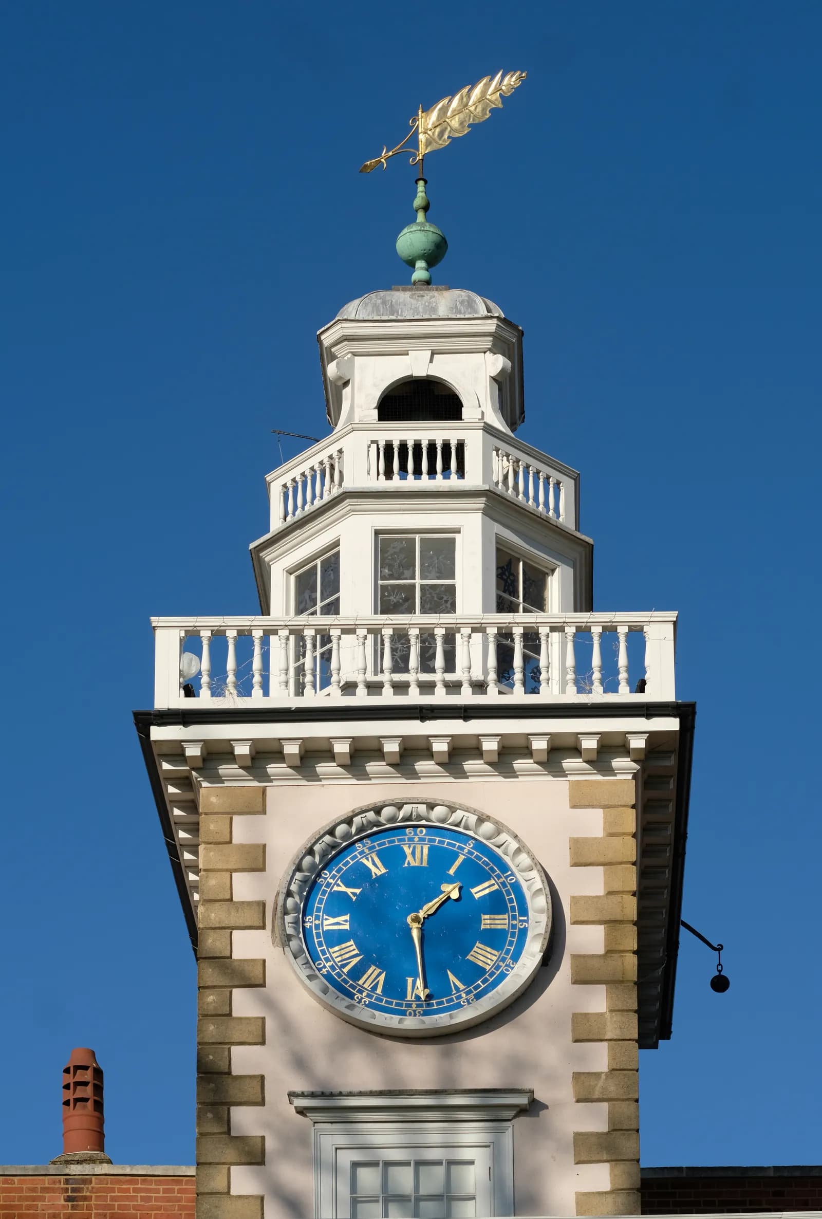 A close-up view of a detailed clock tower with a golden leaf-shaped weather vane on top, featuring a striking blue clock face with gold Roman numerals and hands.