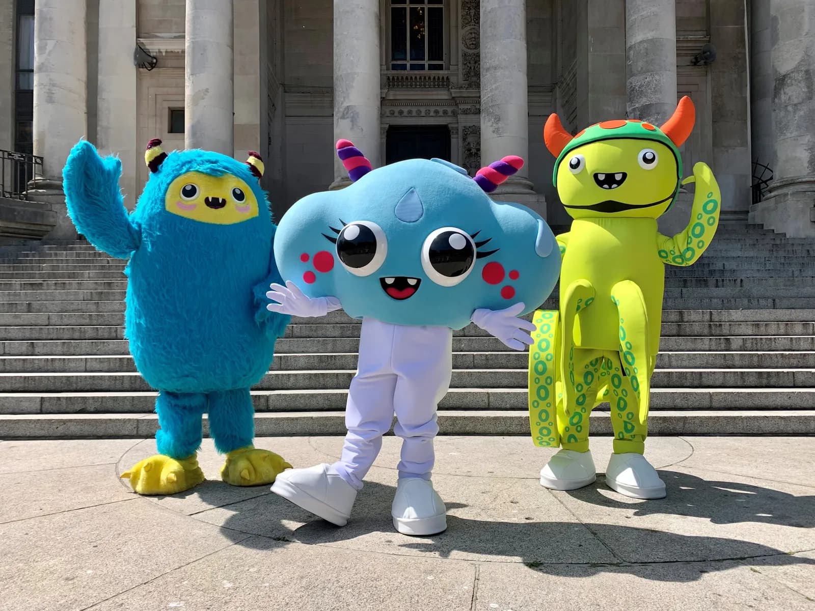 Three colorful mascot costumes, a blue furry monster, a light blue cloud-like figure, and a green monster with horns, stand outdoors in front of a building with large columns and stone steps.