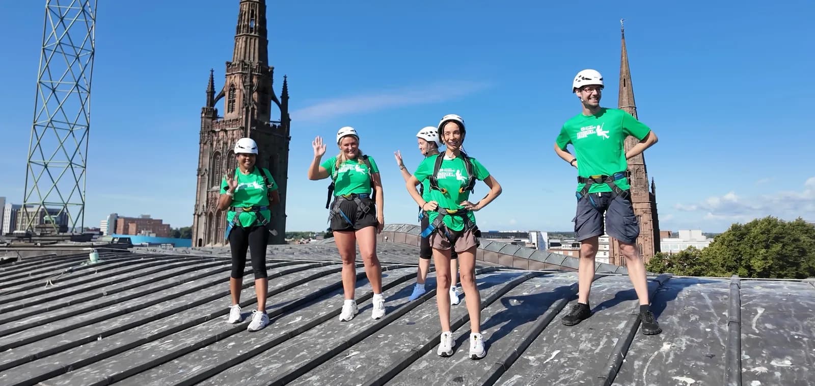 Five people in green shirts, white helmets, and harnesses pose on a rooftop with a tall, gothic-style stone tower in the background under a clear blue sky.
