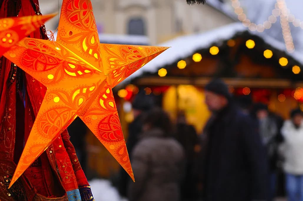 A close-up of a decorative orange star-shaped lantern with intricate patterns, illuminated and hanging, with a blurred background of a bustling winter market with people and lights.