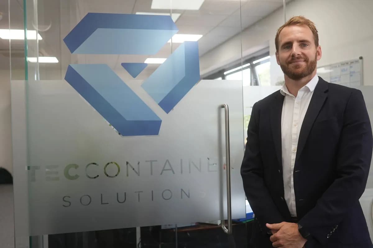A man in a navy suit and white shirt smiles while standing in front of a glass door with a company logo and name printed on it.