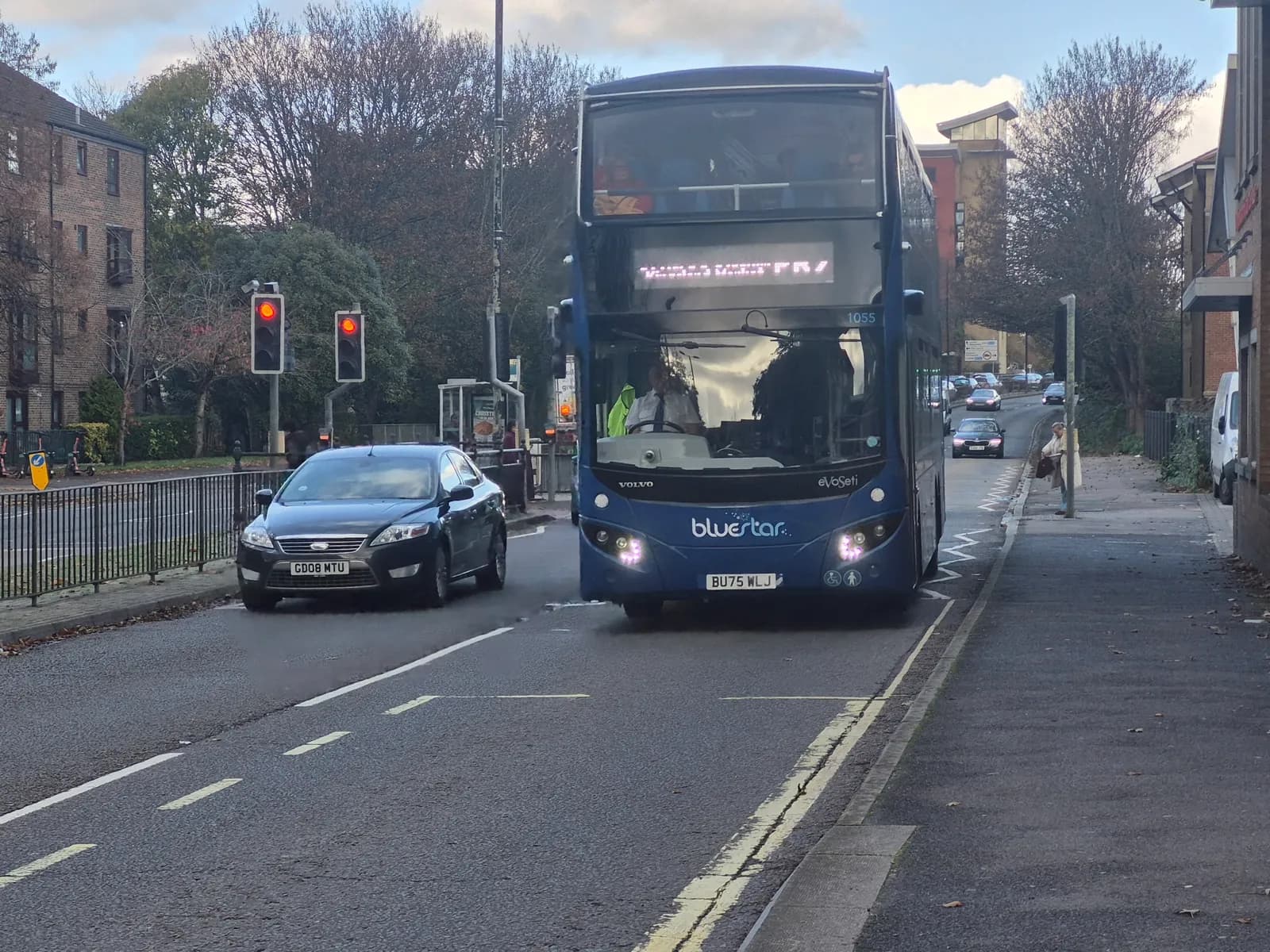 A blue double-decker bus from Bluestar is stopped in traffic next to a black car, with traffic lights showing red in the background.