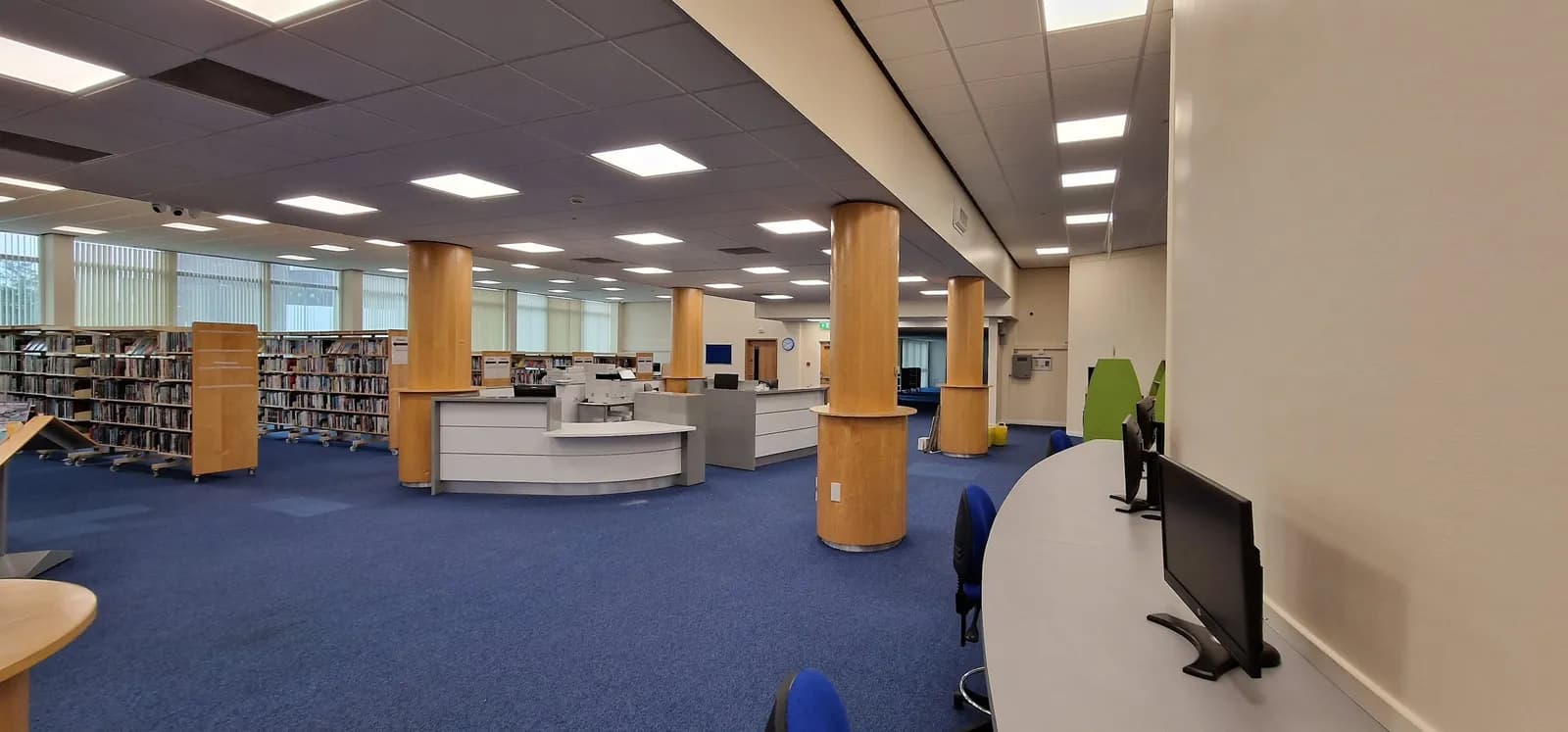 A wide interior shot of a library with blue carpet, bookshelves lining the walls, and several light-colored service desks and tall columns in the center.