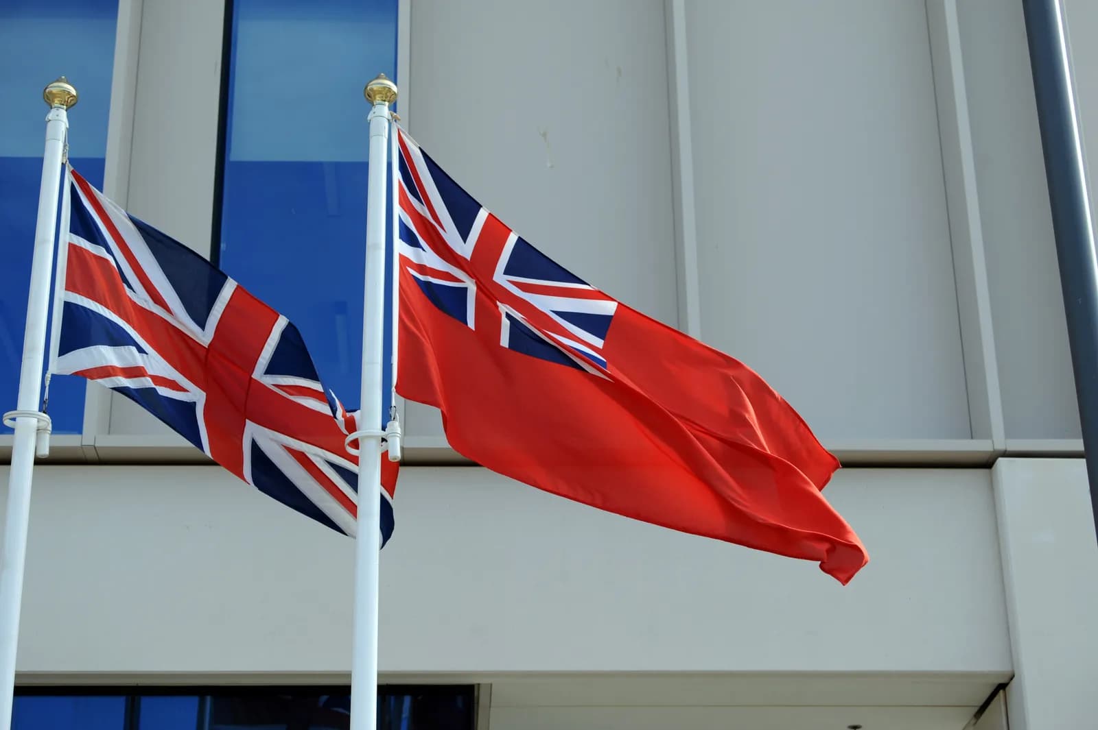 Two Red Ensign flags, each featuring a Union Jack in the upper left canton, wave from white flagpoles against a modern building with blue-tinted windows.