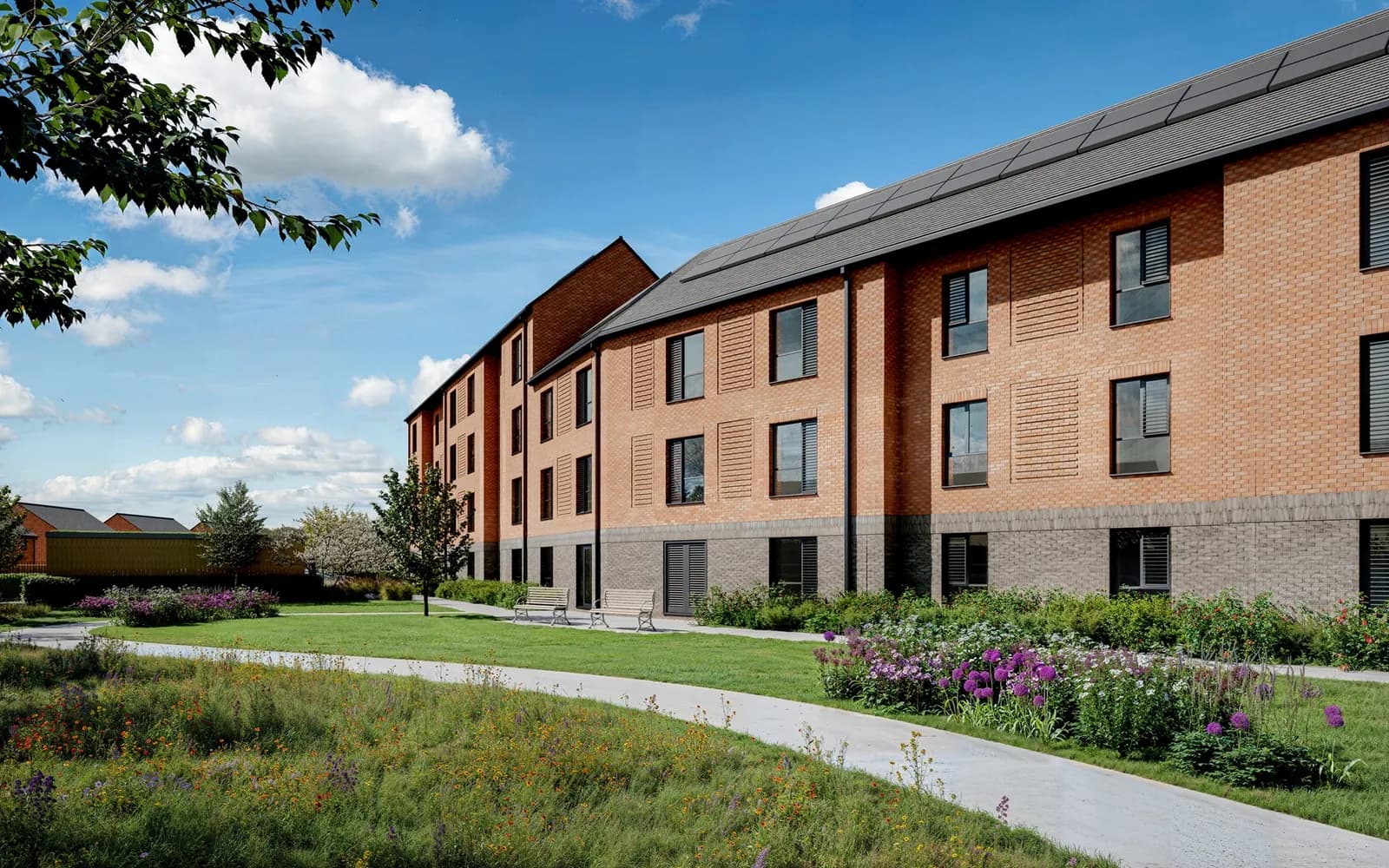 A modern brick building with solar panels on the roof and neatly landscaped gardens with a path in front.