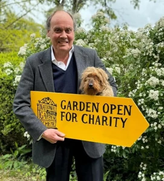 A man in a suit jacket holding a yellow directional sign that reads "GARDEN OPEN FOR CHARITY" with a small, shaggy brown dog looking over the sign, all set against a backdrop of green foliage with white flowers.