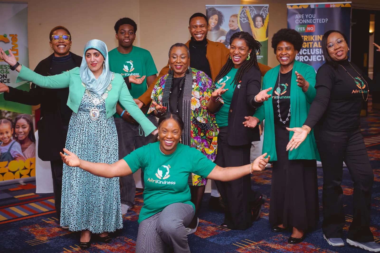 A group of ten diverse individuals smile and pose with outstretched arms, some wearing green t-shirts with an "AfriKindness" logo.