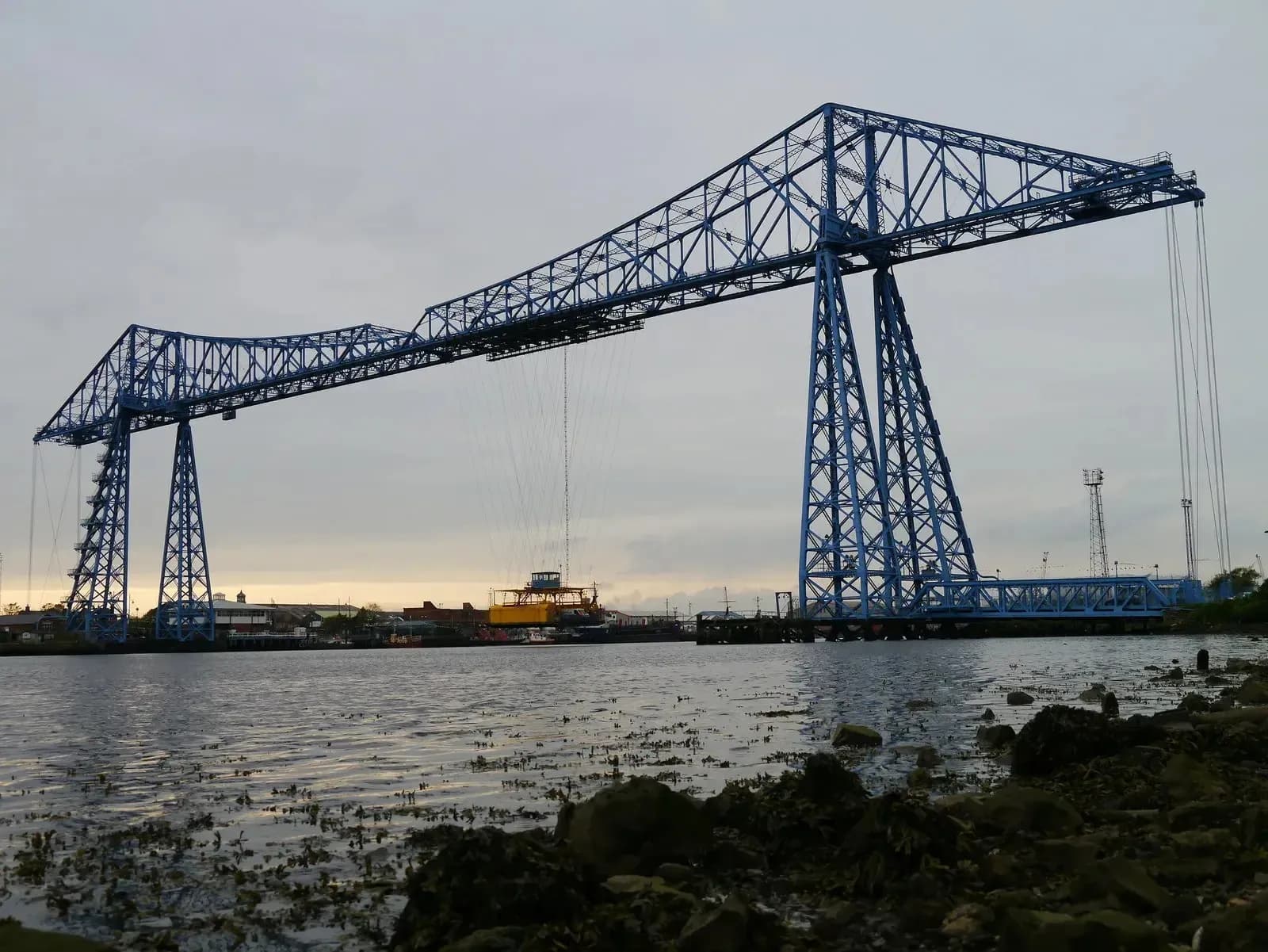 A large blue transporter bridge spans across a wide body of water, with a yellow gondola suspended below its main structure, under a cloudy sky.