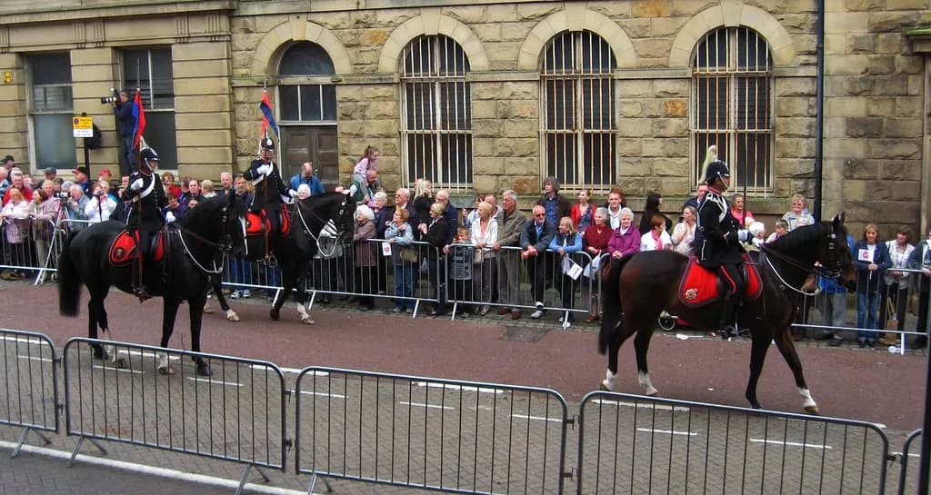 A parade with several mounted cavalry officers in dark uniforms with red detailing, riding black and brown horses, passing by a crowd of spectators behind metal barriers, in front of a stone building with arched windows.