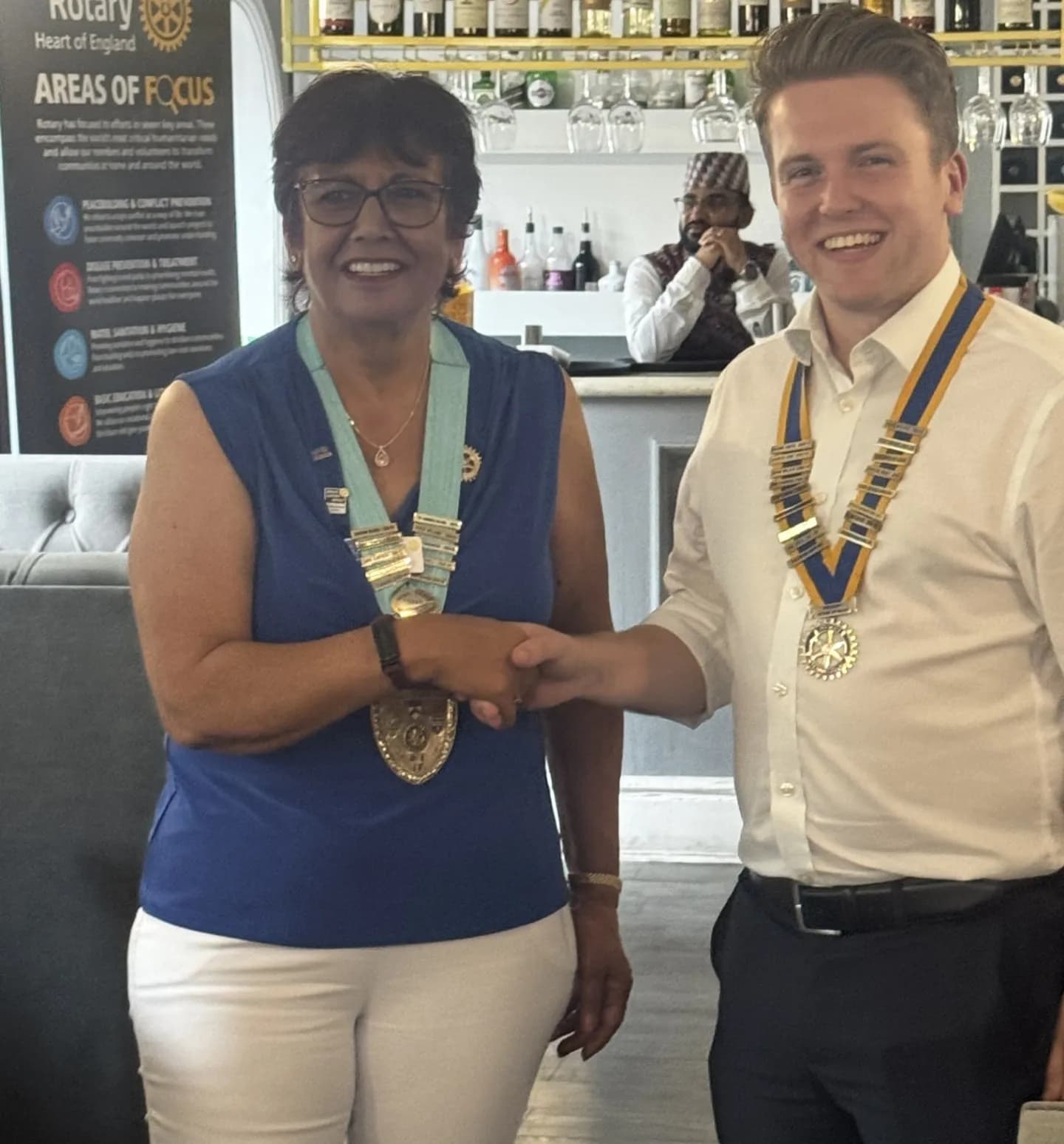 A woman and a man, both wearing blue and gold sashes with medallions, shake hands and smile in a brightly lit room with a bar and a Rotary banner in the background.