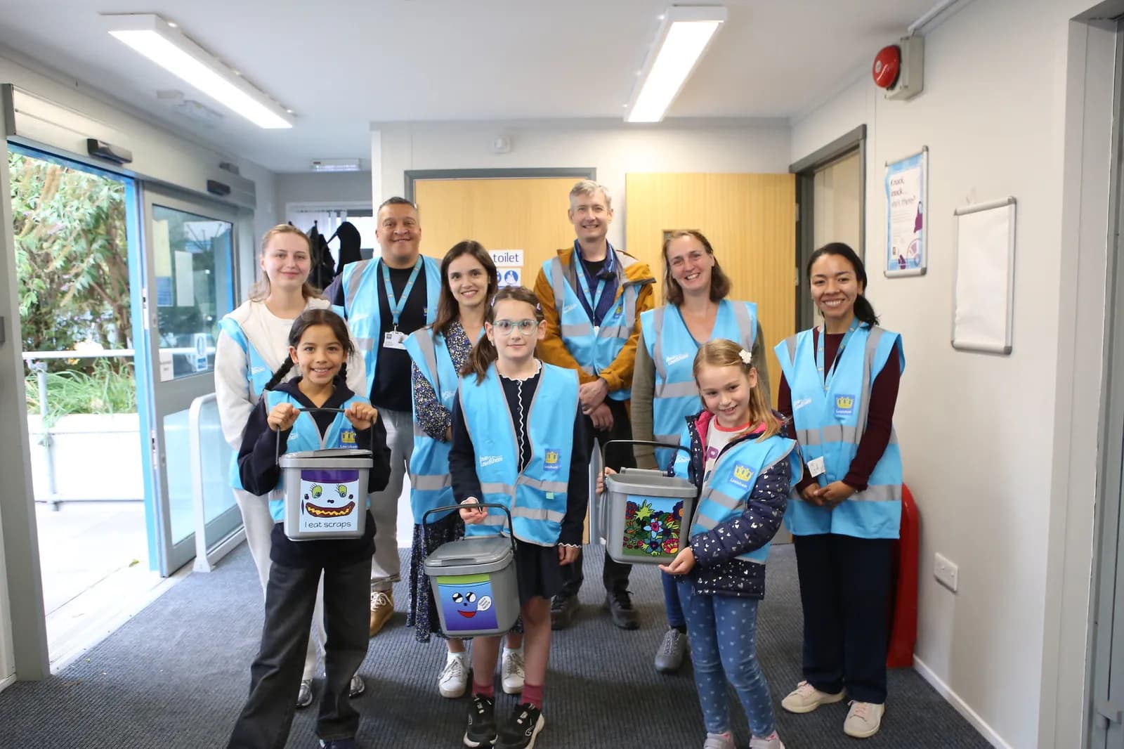 A group of adults and children wearing light blue vests and holding donation buckets stand in a hallway.