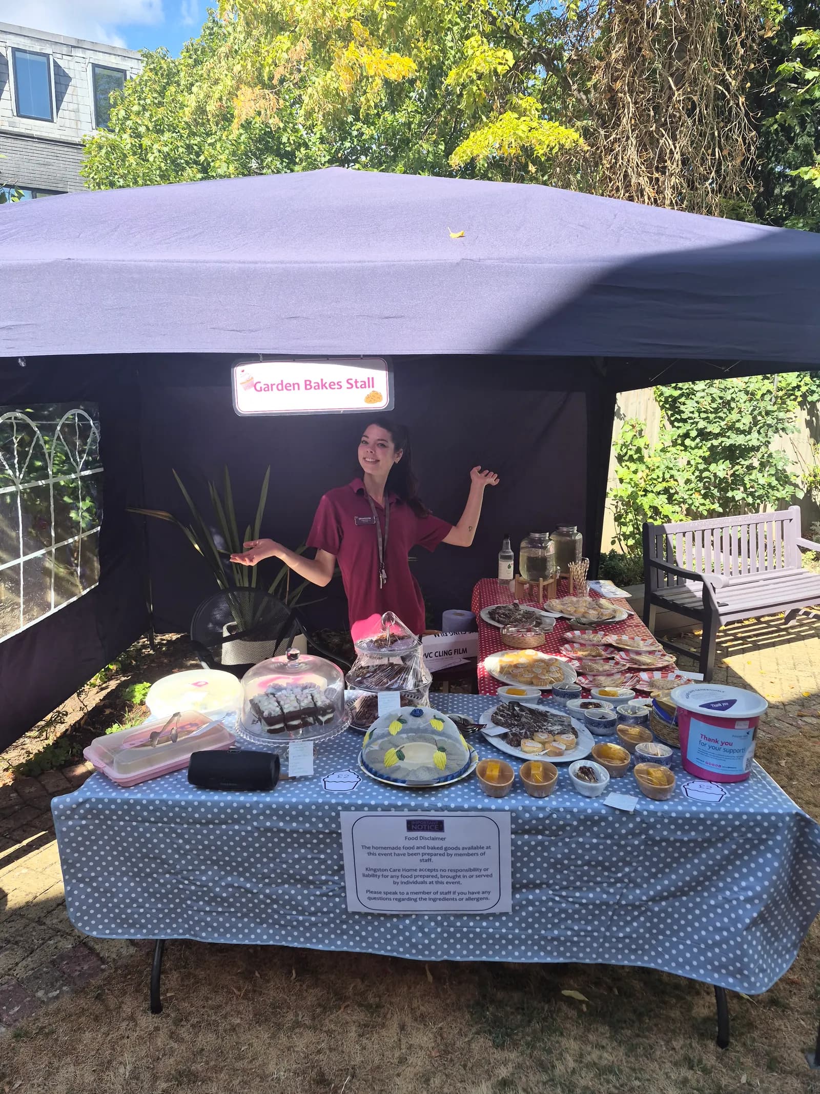 A smiling woman stands behind a table filled with baked goods and drinks under a purple tent, at a 'Garden Bakes Stall'.