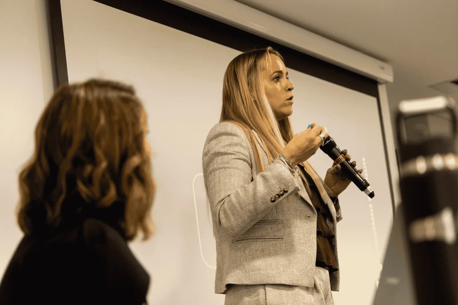 A woman with long blonde hair, wearing a grey tweed suit, holds a microphone and speaks while another person with brown curly hair looks on from the left.