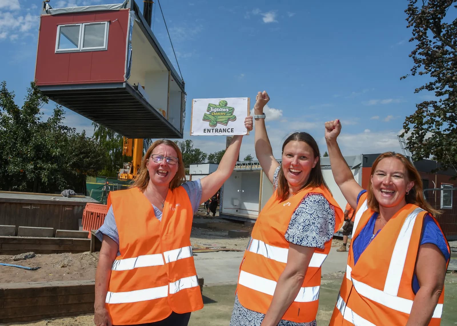 Three women in orange vests celebrating the placement of a new modular building, with one holding a sign that says "Jigsaws Childcare Entrance."