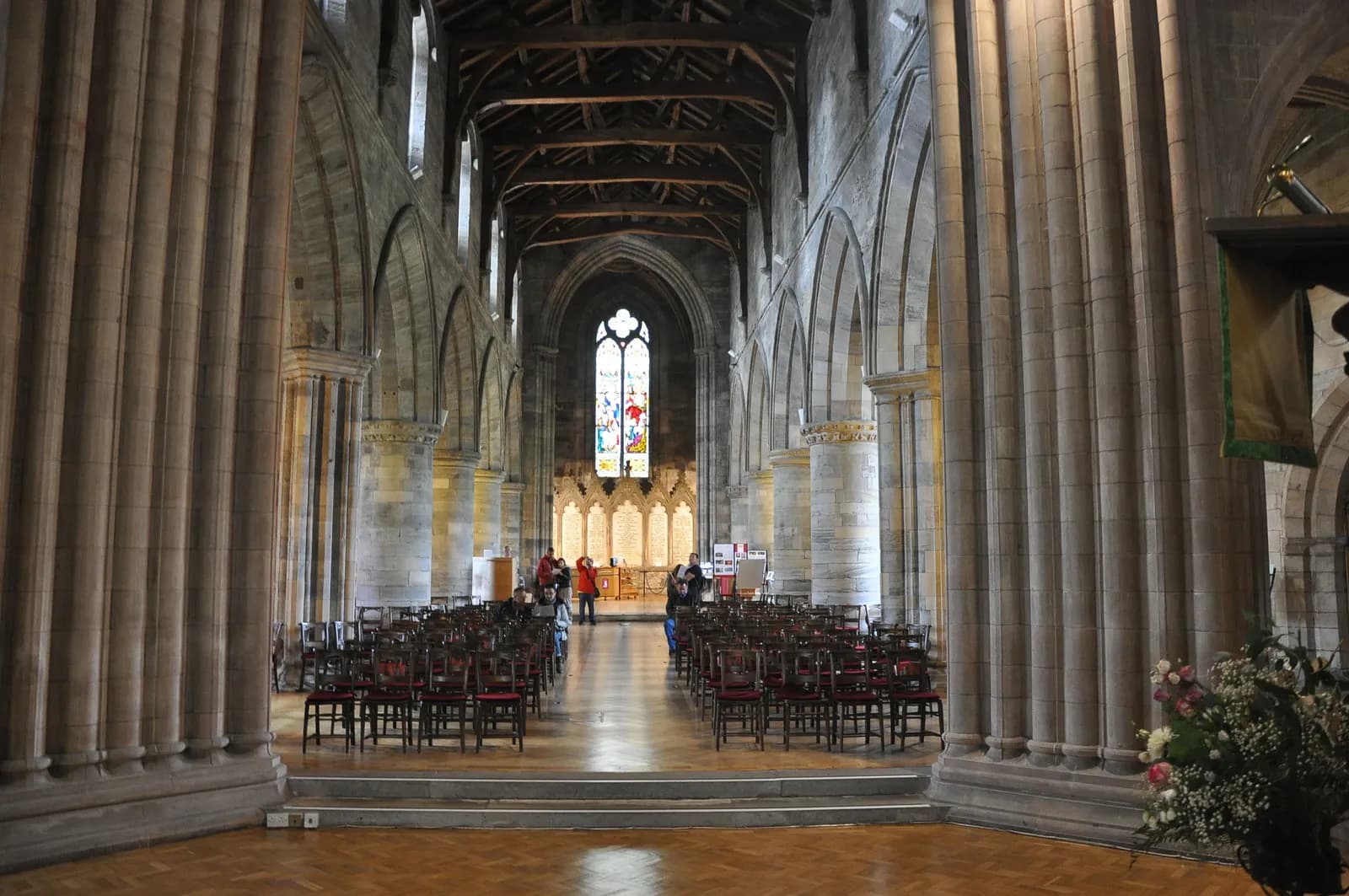 The interior of a large cathedral with high vaulted ceilings, large stone columns, and rows of empty wooden chairs leading towards a colorful stained-glass window in the distance.