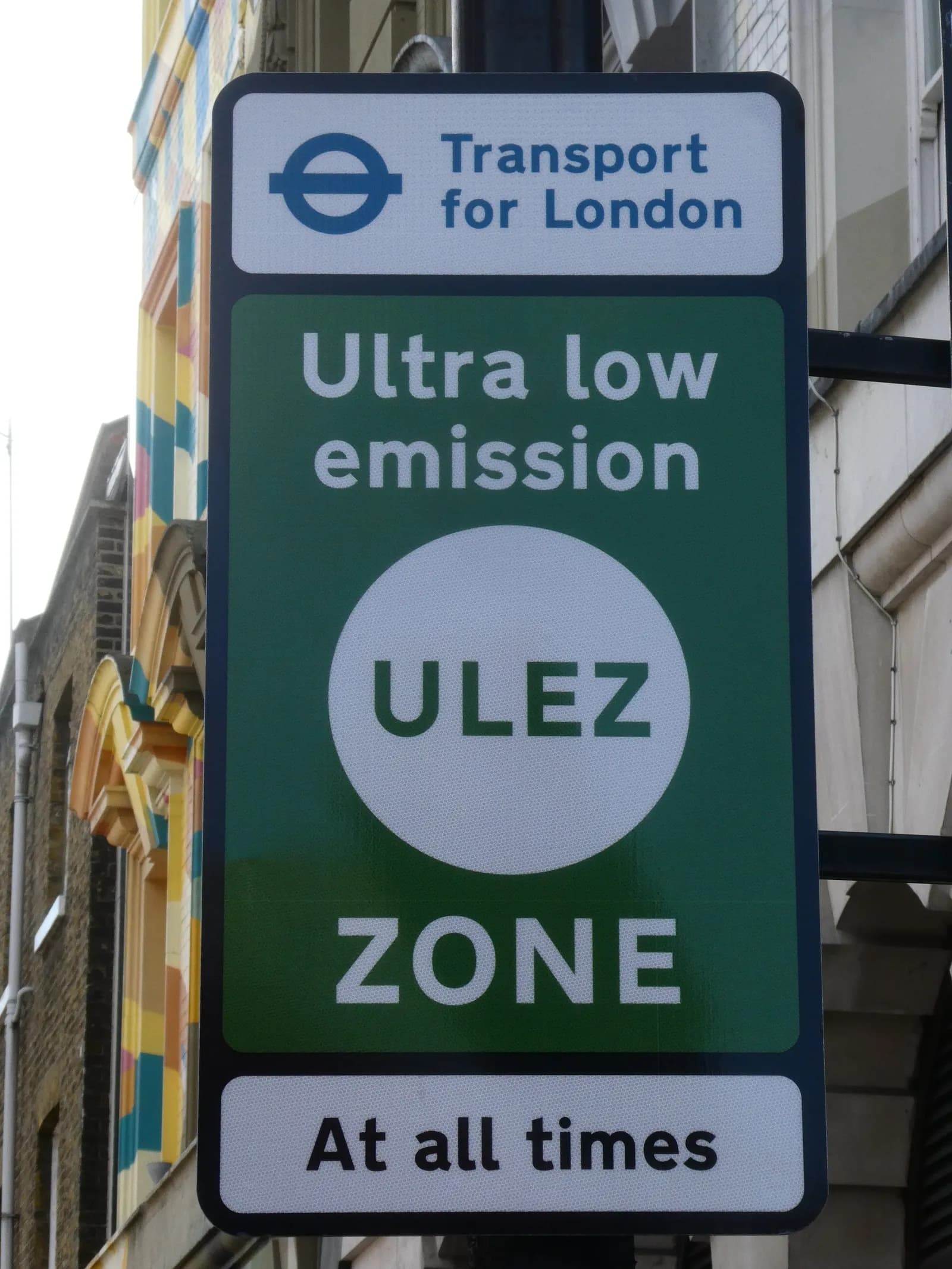 A green and white sign for the Transport for London Ultra Low Emission Zone (ULEZ) hangs on a pole with a building in the blurred background.