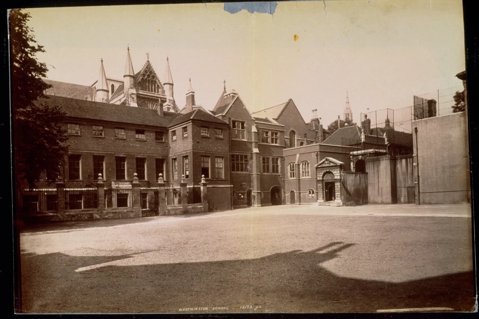 A sepia-toned photograph shows the old stone and brick buildings of Westminster School surrounding a graveled courtyard, with a tree on the left and the spires of Westminster Abbey visible in the background against a light sky.