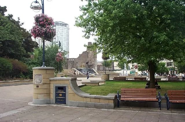 A park scene with a large tree in the foreground, benches, a fountain, and a ruined building in the background.