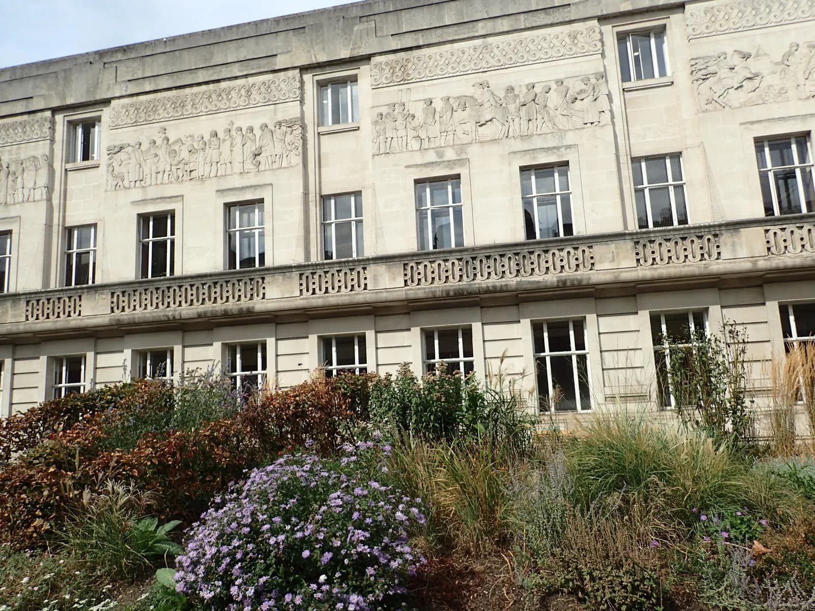 A large stone building with multiple windows, decorative balustrades, and elaborate historical carvings on its facade, partially obscured by a vibrant foreground of green and purple foliage.