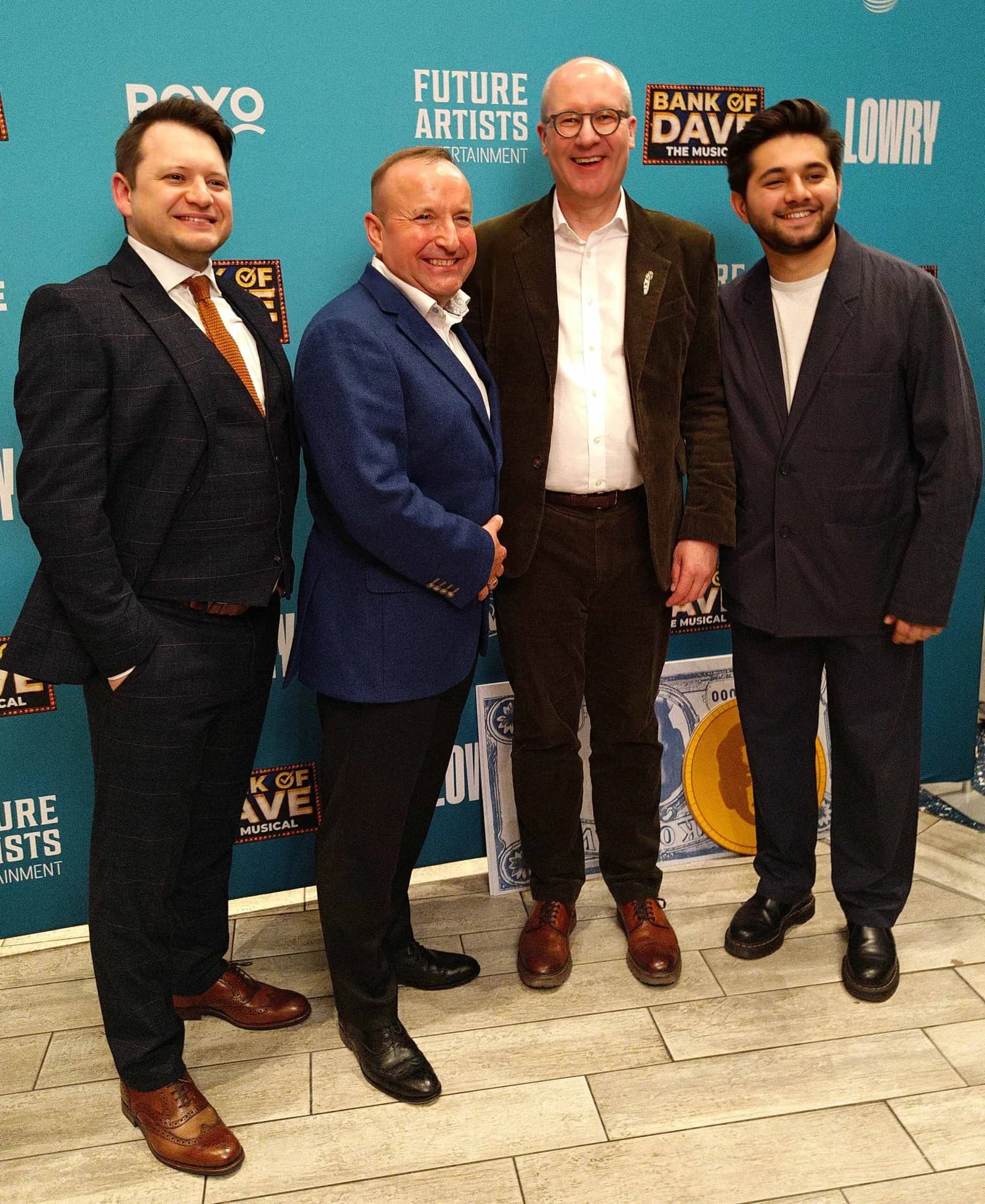 Four well-dressed men smiling at the camera in front of a blue backdrop with event branding.