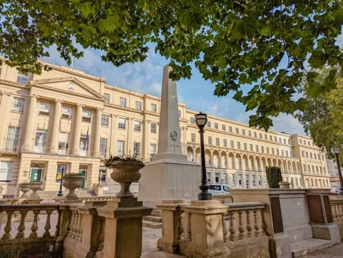 A large, light-colored neoclassical building with columns and many windows is framed by green tree leaves at the top, with a tall obelisk-like monument and lamppost in the foreground.