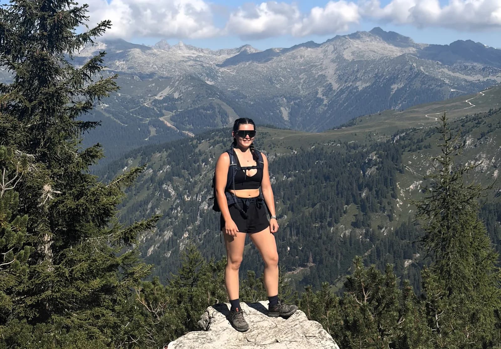 A woman wearing sunglasses, a black sports bra, black shorts, and a backpack stands on a rock with a mountainous landscape and forests in the background.