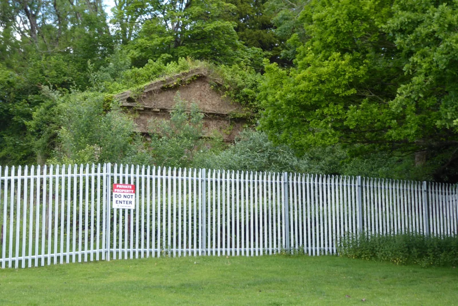 A white fence with a red "Private Property Do Not Enter" sign in front of a heavily overgrown building surrounded by lush green trees.