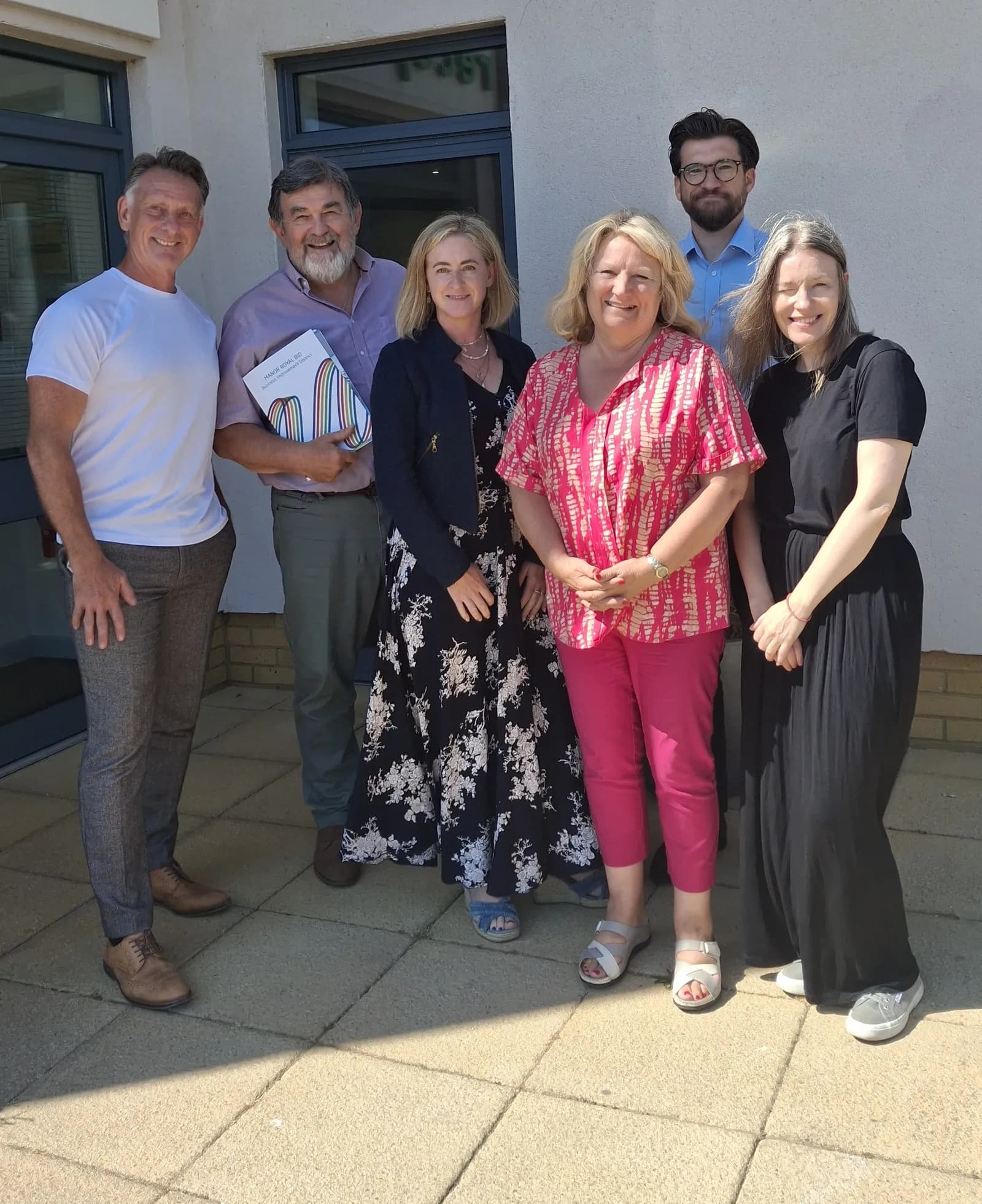 Six people, four women and two men, stand smiling together outside a building on a sunny day.