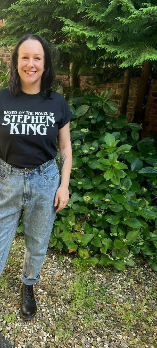 A smiling woman wearing a black t-shirt with “BASED ON THE NOVEL BY STEPHEN KING” printed on it and light blue jeans stands in a garden.