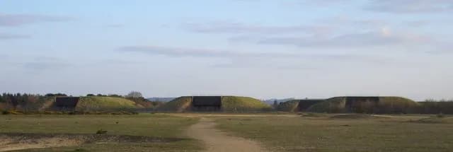 A wide shot of a field with four grass-covered bunkers, each featuring a dark, rectangular opening, under a partly cloudy sky.