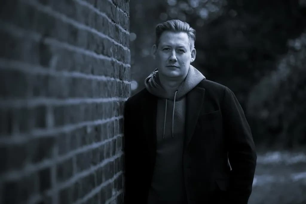 A man with slicked-back hair and a light hoodie under a dark jacket leans against a brick wall, looking directly at the viewer.