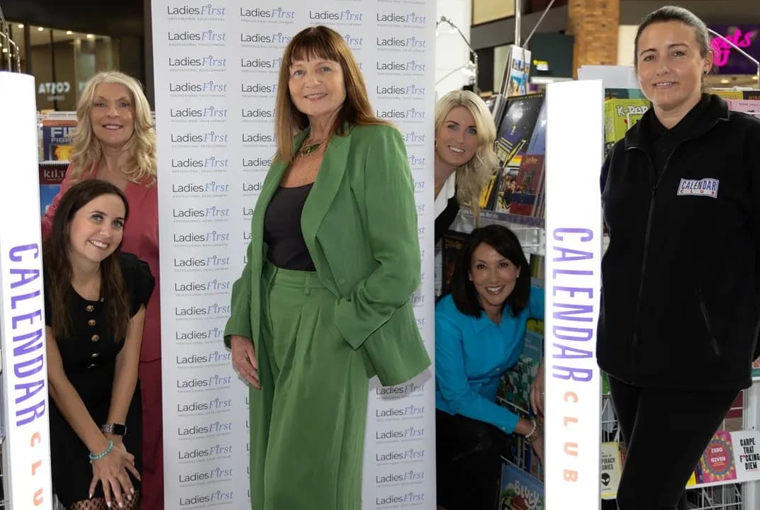 A group of six women, one in a green pantsuit, gathered around a "Ladies First" banner and "Calendar Club" displays.