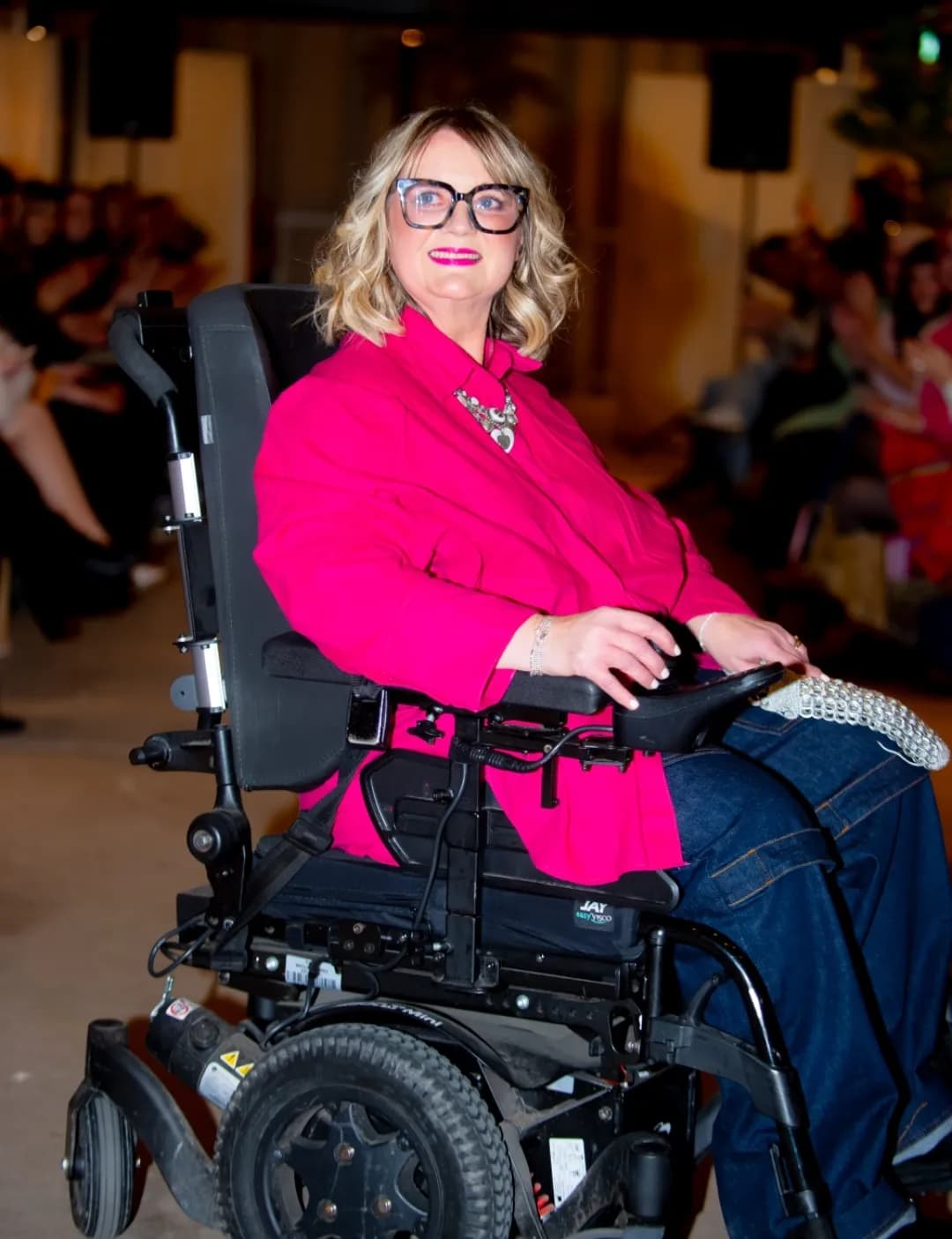 A woman with blonde wavy hair and glasses smiles while sitting in a motorized wheelchair and wearing a bright pink top, dark blue jeans, and a silver necklace.