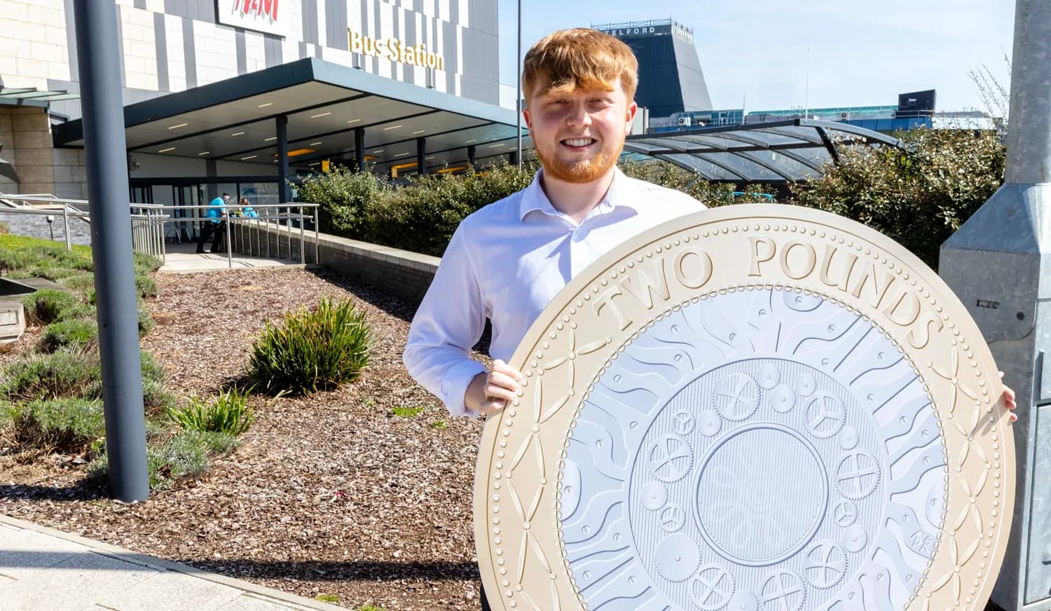 A smiling young man with red hair and a white shirt holds a giant two pounds coin in front of a bus station.