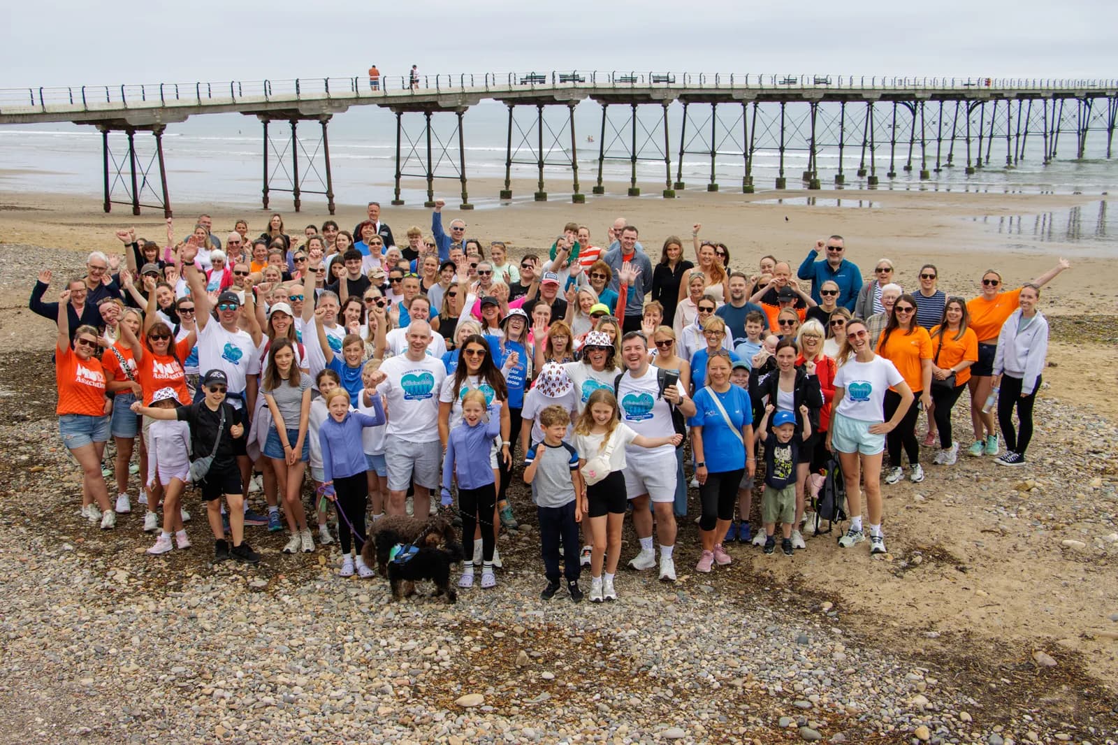 A large group of people wearing blue, orange, and white t-shirts are standing and smiling on a rocky beach, with a long pier stretching over the water in the background.