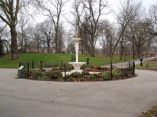 A white decorative fountain is enclosed by a black metal fence in the center of a park with bare trees and green grass under an overcast sky.