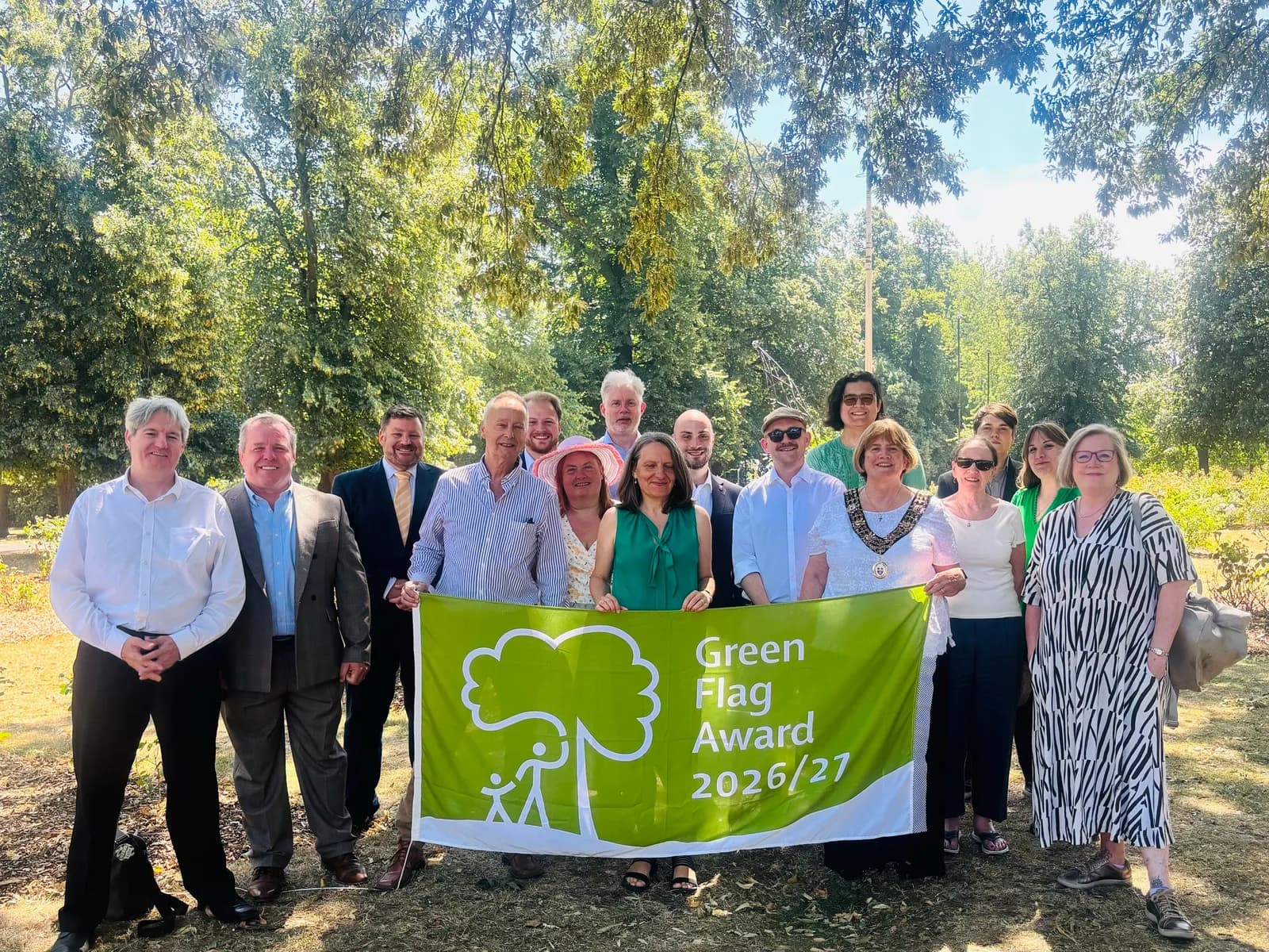 A group of people, including women and men, stand outdoors under trees, holding a green banner that reads "Green Flag Award 2026/27."