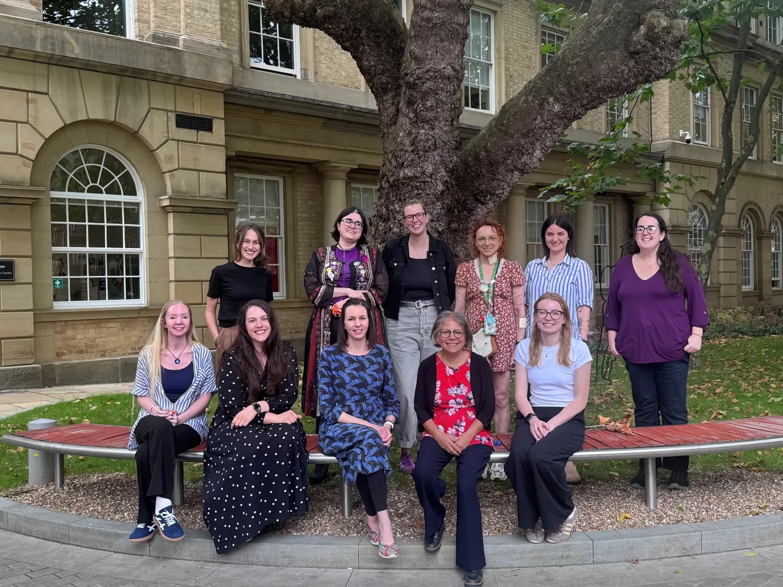 A group of ten smiling women in casual attire pose outdoors in front of a historic stone building with a large tree.