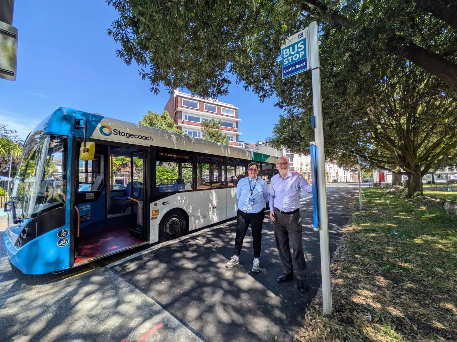 A man and a woman stand smiling next to a blue Stagecoach bus with an open door, next to a bus stop sign that reads "BUS STOP Castle Road".