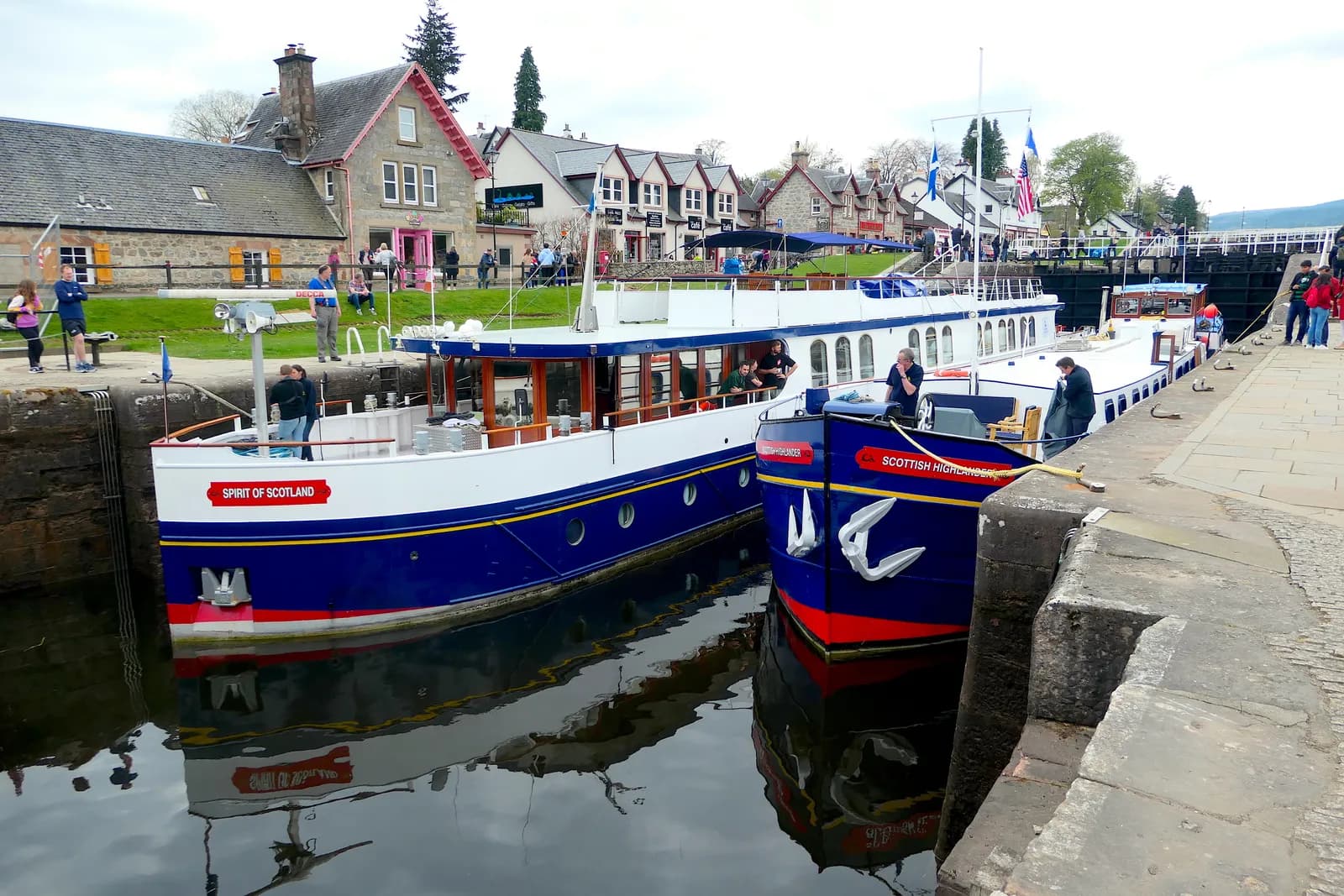 Two boats, 'Spirit of Scotland' and 'Scottish Highlander,' are docked on a canal with several buildings and people in the background.