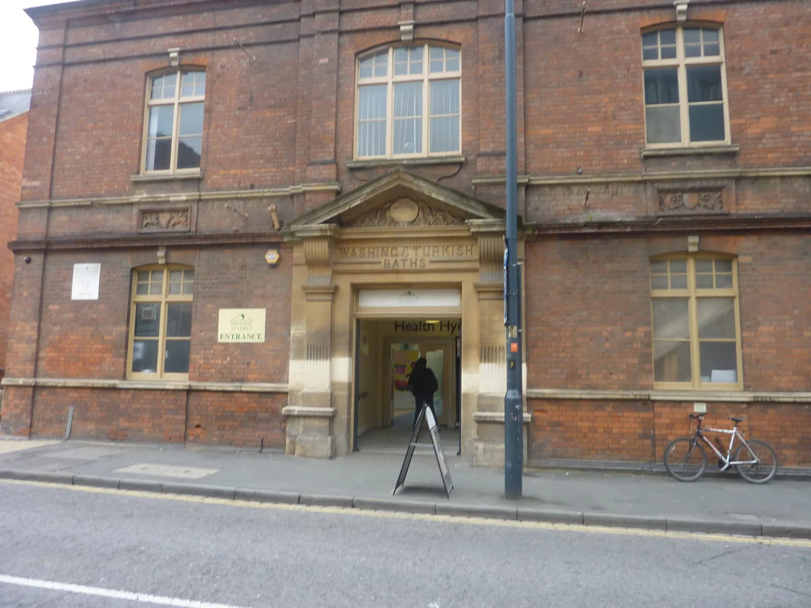 The brick facade of a historic building with large arched windows and an ornate stone entrance to the Washing and Turkish Baths, with a person walking into the entrance and a bicycle parked on the sidewalk to the right.