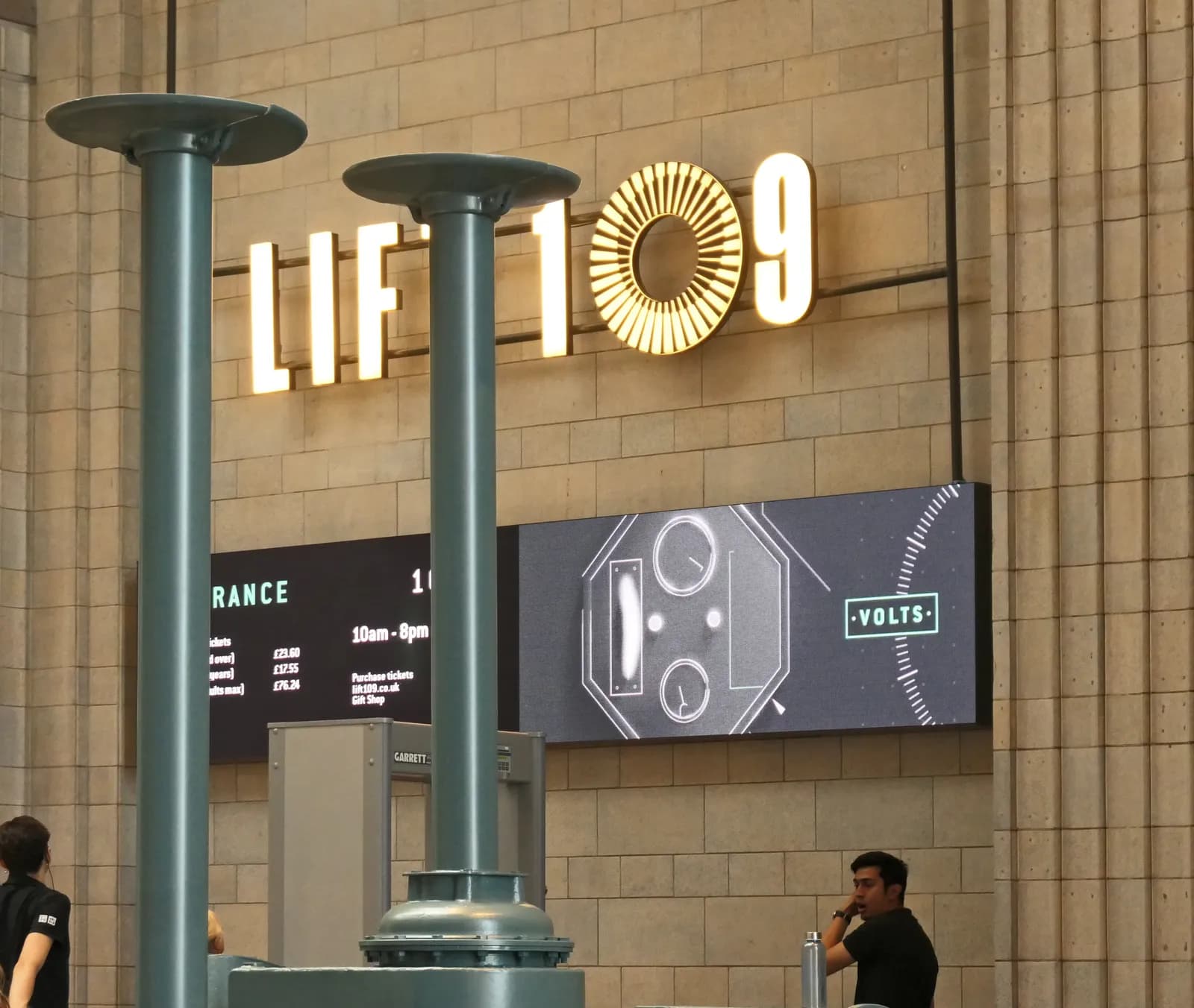 A large illuminated sign for "LIFT 109" above two electronic display boards and two people, one of whom is talking on the phone.