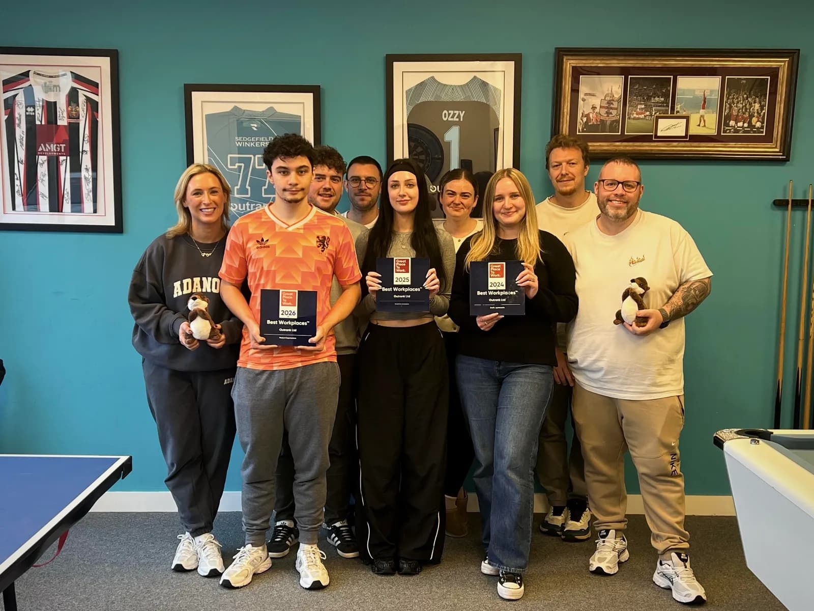 A diverse group of ten people, some holding framed awards and stuffed animals, pose together in a room with framed jerseys, photos, and a pool table.