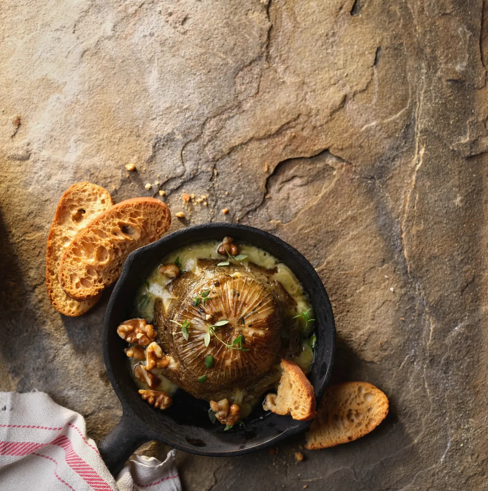 A baked onion with herbs and cheese sauce in a black skillet, served with sliced toasted bread and walnuts on a stone surface.