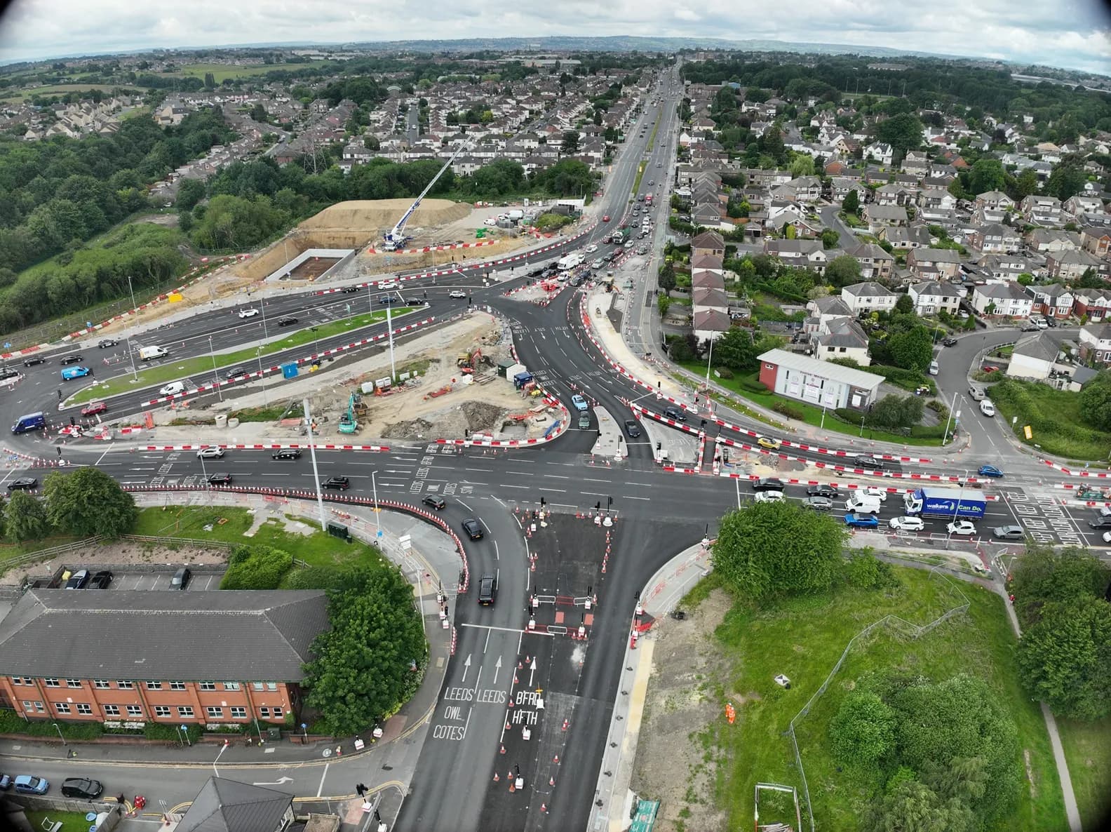 An aerial view shows a large road construction project with multiple lanes, ongoing earthworks, and surrounding residential areas.