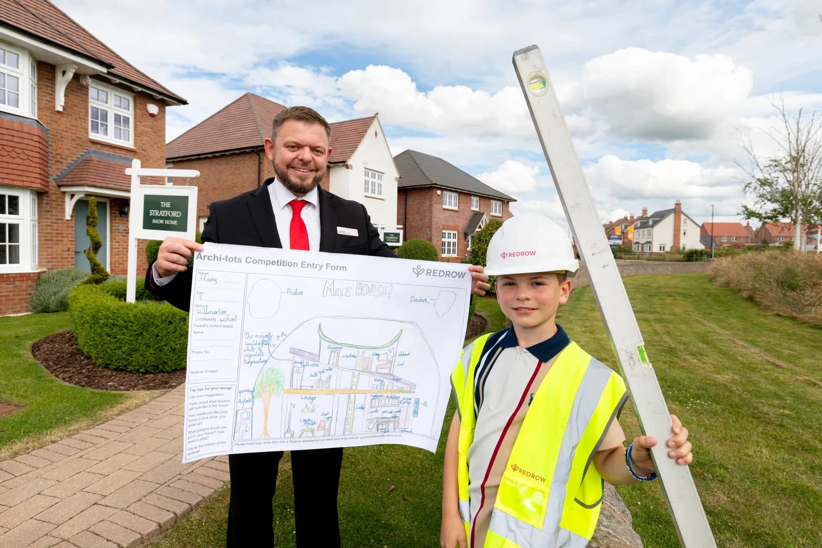 A man in a suit and a young boy in a construction vest and hard hat stand outdoors, holding a competition entry form and a spirit level, in front of residential homes.