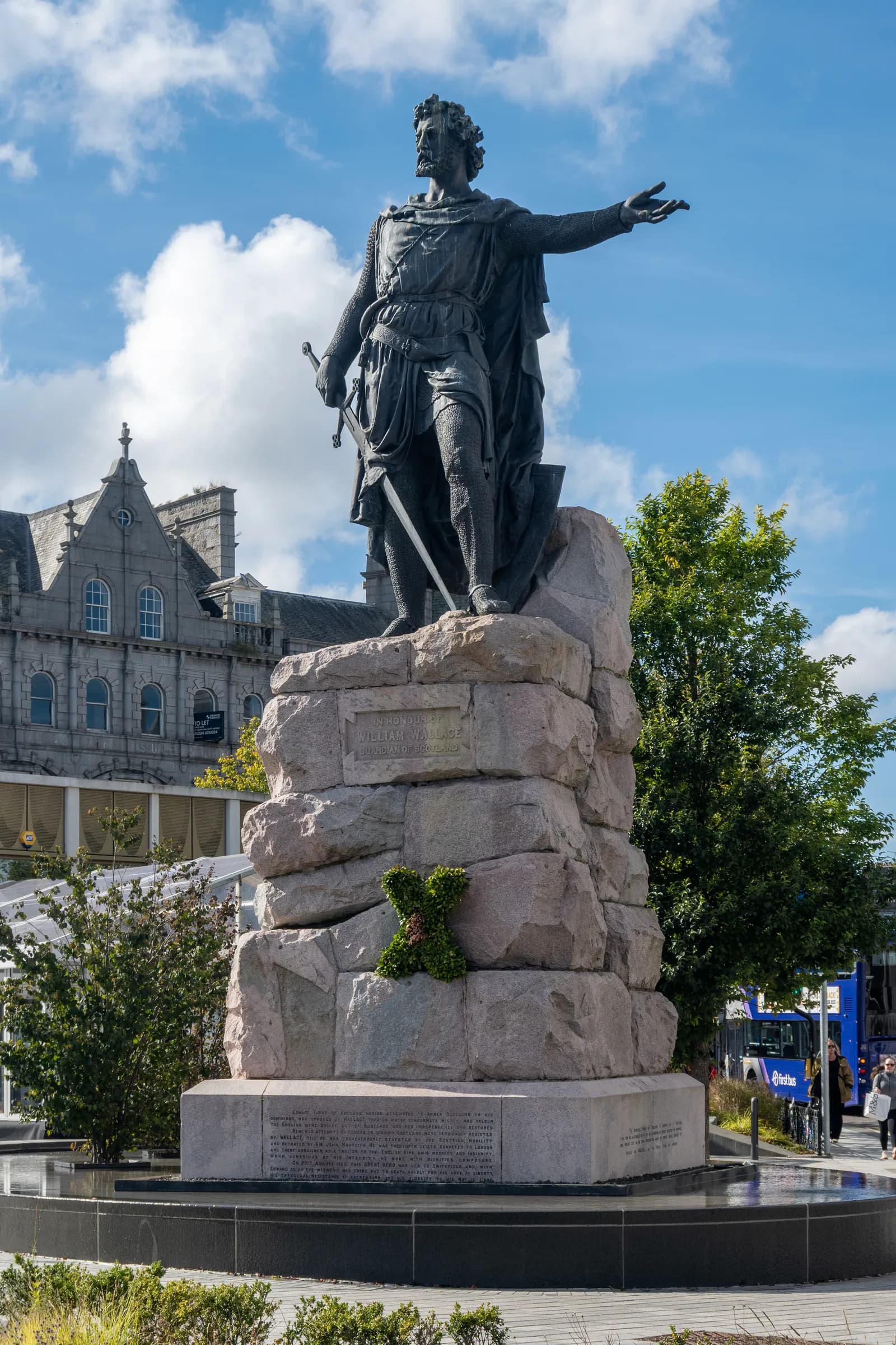 A large bronze statue of a man in armor holding a sword and pointing, standing on a stone pedestal against a blue sky with clouds and a grey building in the background.