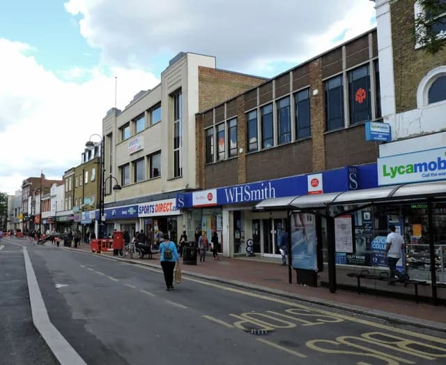 A bustling street scene features a row of shops on the right, including WHSmith and Sports Direct, with pedestrians and a bus stop.